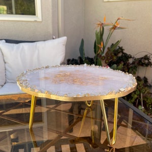 May include: A white and gold resin cake stand with a gold metal base. The stand is decorated with small clear crystals around the edge.