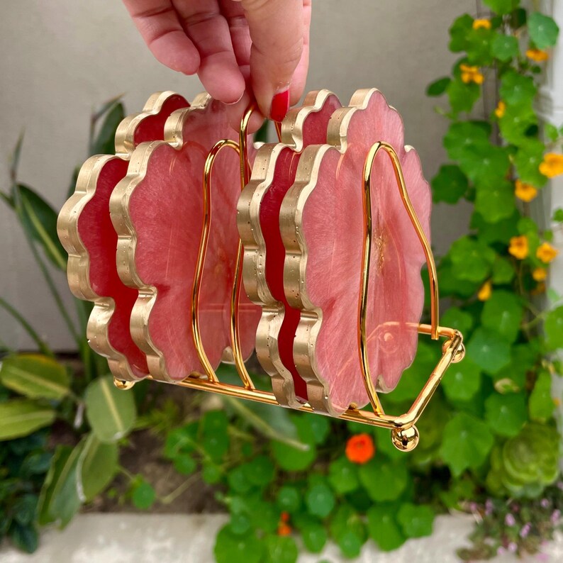 Puede incluir: Conjunto de cuatro posavasos de resina rosa y dorada con borde festoneado, presentados en un soporte de metal dorado.