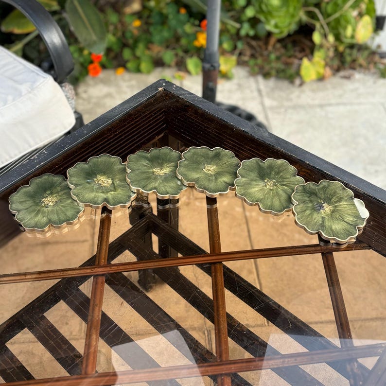 Peut inclure: Ensemble de six sous-verres en r&eacute;sine verte et dor&eacute;e avec un motif floral. Les sous-verres sont dispos&eacute;s en demi-cercle sur une table en verre avec un cadre en bois.