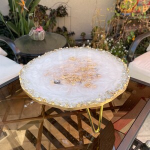 May include: White resin serving tray with gold flecks and a gold metal stand. The tray has a round, irregular shape and is decorated with small, clear crystals around the edge.