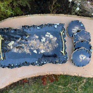 May include: A black tray with gold flecks and a gold trim, along with four matching coasters. The tray and coasters have a unique, irregular shape.
