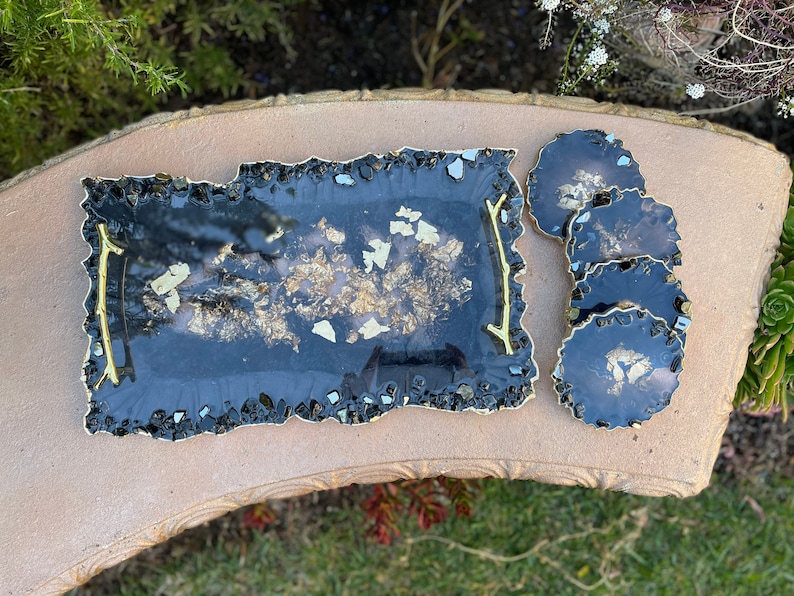 May include: A black tray with gold flecks and a gold trim, along with four matching coasters. The tray and coasters have a unique, irregular shape.