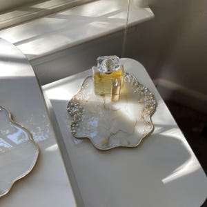 May include: A decorative white and gold resin tray with a floral design, holding a perfume bottle and a small vial. The tray has a gold trim and is adorned with pearl-like embellishments. The perfume bottle is clear with a gold cap.