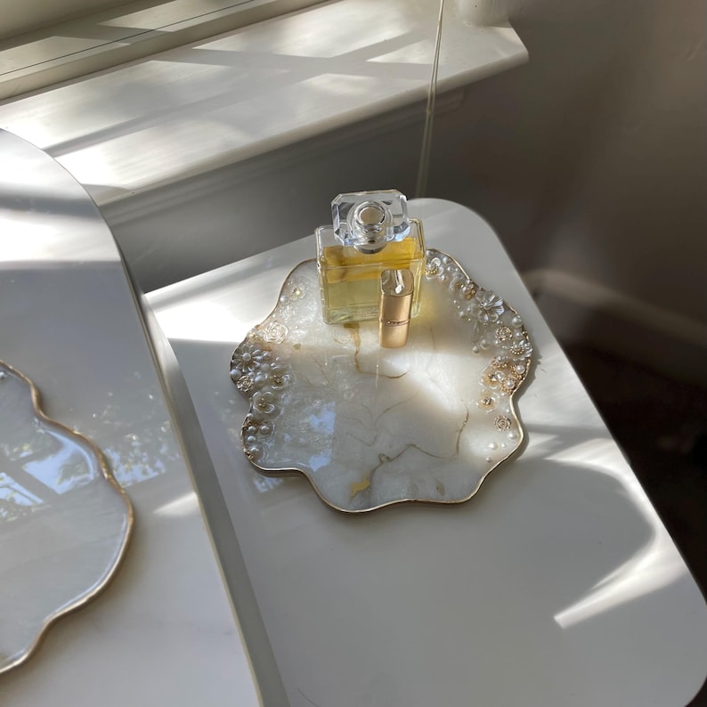 May include: A decorative white and gold resin tray with a floral design, holding a perfume bottle and a small vial. The tray has a gold trim and is adorned with pearl-like embellishments. The perfume bottle is clear with a gold cap.