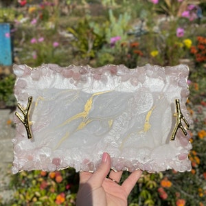 May include: White resin tray with gold accents and a pink quartz border. The tray has two gold metal handles in the shape of branches.