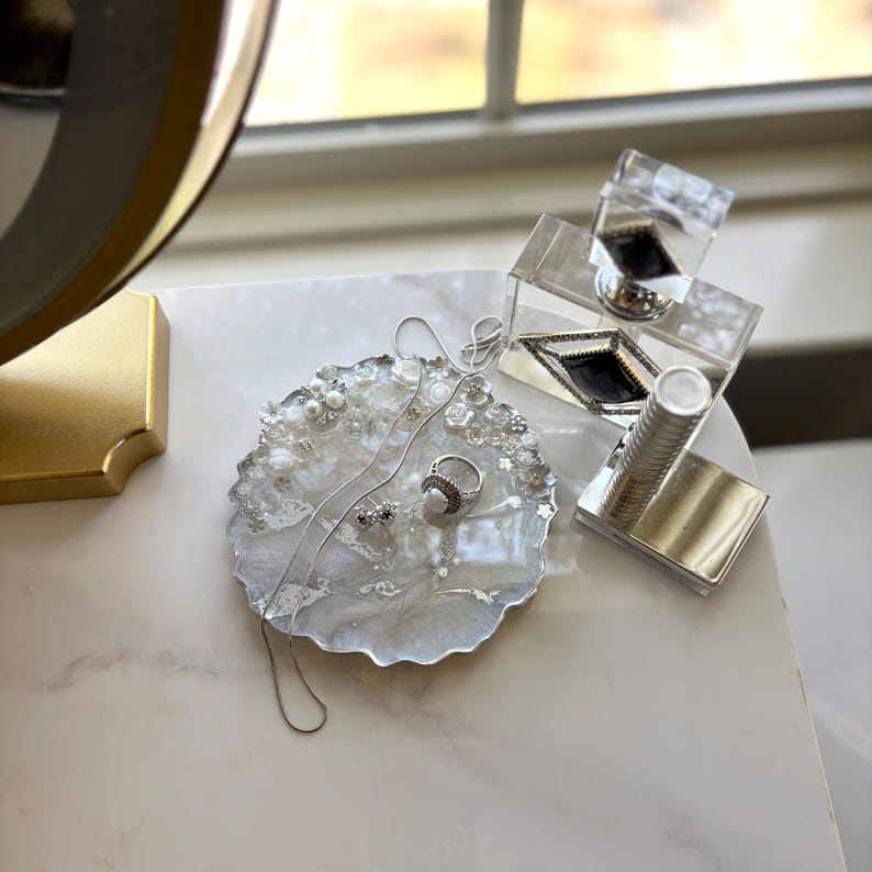 May include: A decorative jewellery tray with a white and grey marbled design, adorned with pearls and small white flowers. A silver necklace, ring, and earrings rest on the tray. A clear acrylic box and a silver lipstick holder are also present.