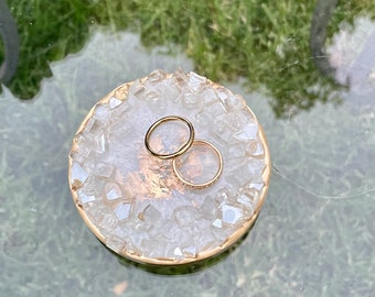 May include: A round, white and gold resin ring dish with a geode-like appearance. Two gold rings rest on the dish. The dish has a gold rim and is on a glass surface with a blurred green background.