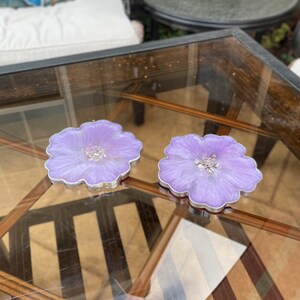 Op de afbeelding: Twee paarse, bloemvormige onderzetters van hars met een zilveren rand. De onderzetters liggen op een glazen tafelblad.