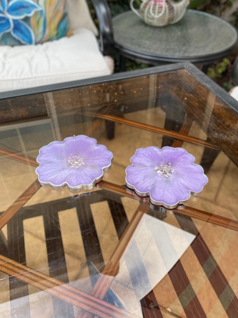 Op de afbeelding: Twee paarse, bloemvormige onderzetters van hars met een zilveren rand. De onderzetters liggen op een glazen tafelblad.