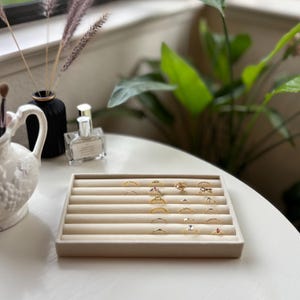 May include: A beige ring display tray filled with various gold rings. The rings feature different designs, including bands, stones, and decorative elements. The tray is on a white table, with a vase, perfume bottle, and plant in the background.
