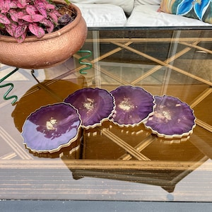 May include: Four purple agate coasters with gold flecks and a gold rim. The coasters are on a glass table with a gold-painted frame.