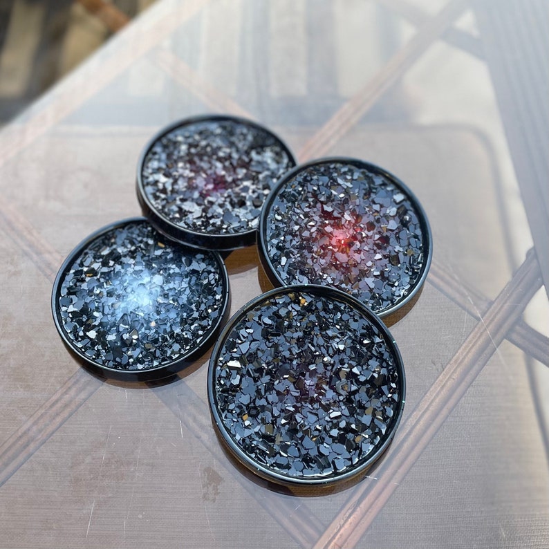 May include: Four black coasters with a glittery finish. Each coaster has a different color of glitter embedded in the resin, including red, blue, and black.