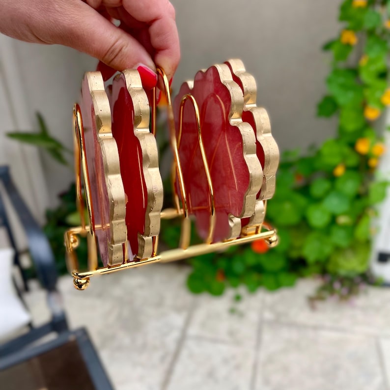 Puede incluir: Conjunto de cuatro posavasos rojos y dorados con borde festoneado. Los posavasos se muestran en un soporte de metal dorado.