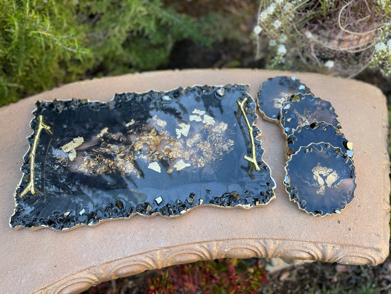 May include: A black and gold resin serving tray with a gold handle and four matching coasters. The tray and coasters have a unique, irregular shape and are decorated with gold flakes.