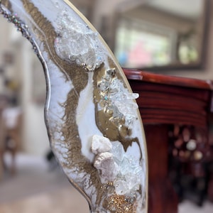 May include: A curved mirror frame featuring a decorative resin pour in white and metallic gold. The surface is embellished with clusters of clear, jagged quartz crystals and fine gold glitter, creating a textured, multi-dimensional appearance.