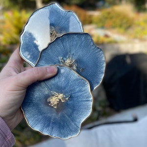 May include: Three blue resin coasters with gold flecks and a white, slightly translucent layer. The coasters are stacked on top of each other and have a slightly irregular, organic shape.