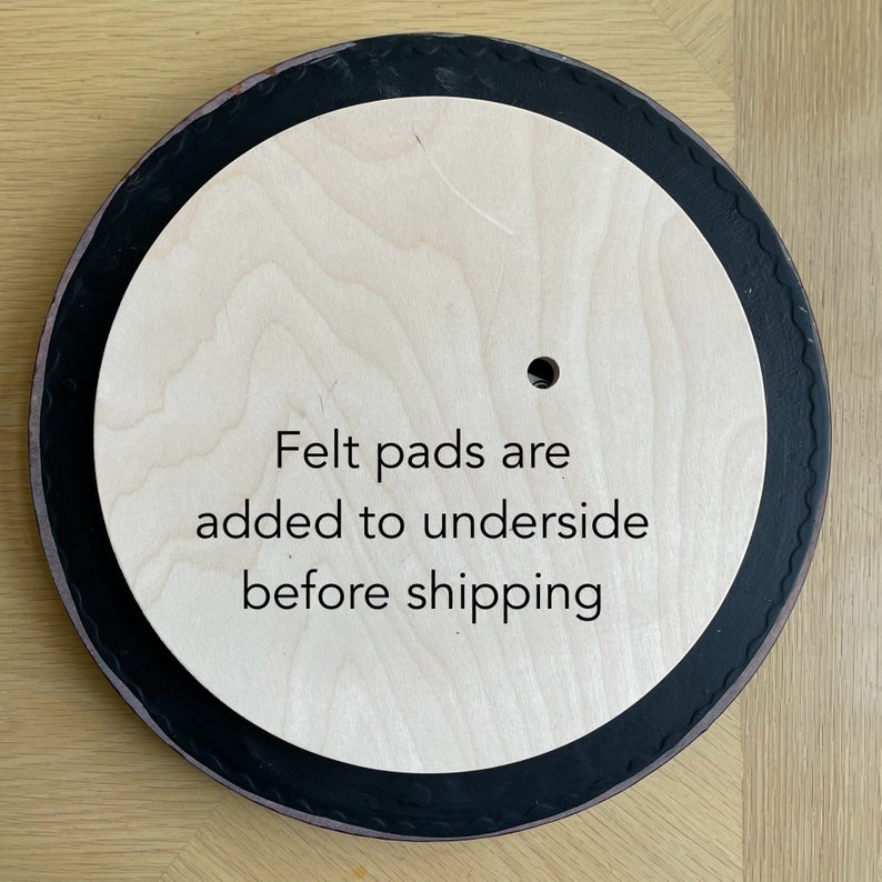 Op de afbeelding: Een ronde houten draaitafel met een zwarte rand. De tekst "Felt pads are added to underside before shipping" is op het oppervlak gedrukt.