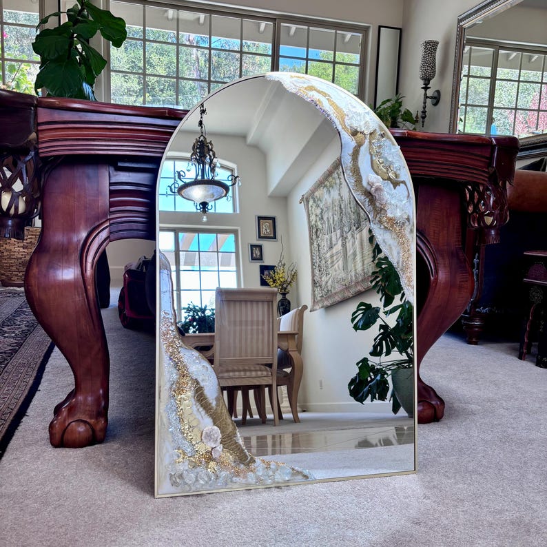 May include: An arched wall mirror featuring decorative resin accents along the top right and bottom left edges. The embellishments consist of textured white, cream, and gold leaf detailing, incorporating embedded clear crystals and stones.