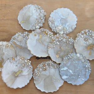 May include: A selection of decorative resin coasters. Each coaster has a unique design with white, gold, and grey swirls, adorned with floral and pearl accents. The coasters are round and vary slightly in size.