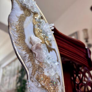 May include: A decorative wall mirror featuring an irregular, organic shape with a white and gold marbled resin finish. The surface is embellished with clusters of clear and pale pink raw crystal points, gold leaf accents, and fine glitter.