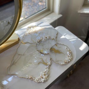 May include: A set of decorative resin serving pieces. The set includes a large, irregularly shaped tray and two smaller coasters. The pieces are white with gold veining, pearl accents, and gold edging. The set is on a white surface.
