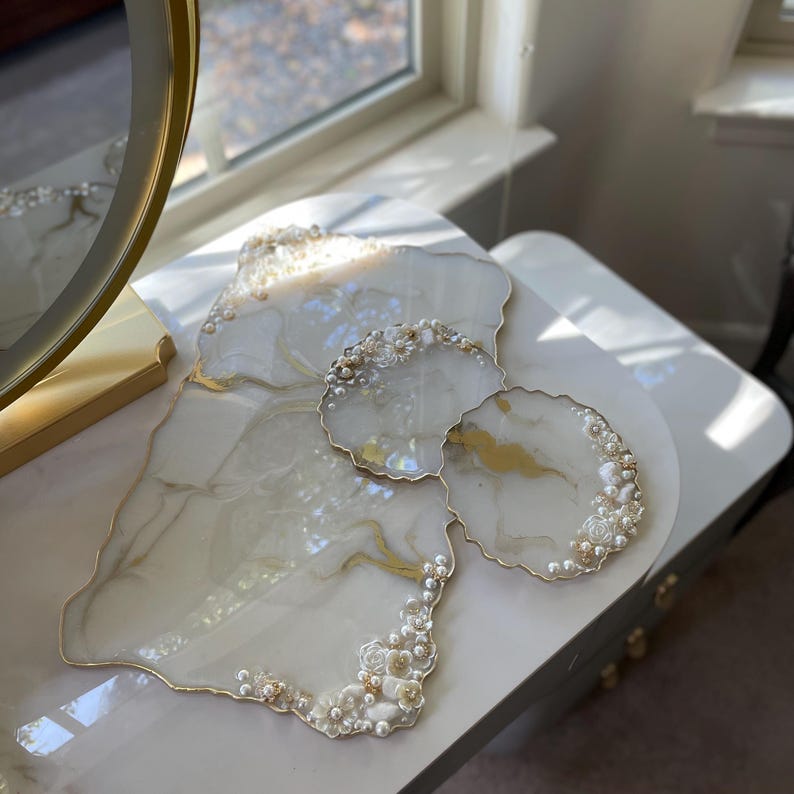 May include: A set of decorative resin serving pieces. The set includes a large, irregularly shaped tray and two smaller coasters. The pieces are white with gold veining, pearl accents, and gold edging. The set is on a white surface.