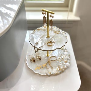 May include: A two-tiered jewelry stand with a gold-colored metal frame. The white resin shelves are decorated with pearls and flowers. The stand holds various necklaces, earrings, and rings.
