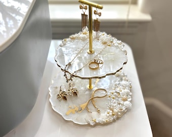May include: A two-tiered jewelry stand with a gold-colored metal frame. The white resin shelves are decorated with pearls and flowers. The stand holds various necklaces, earrings, and rings.