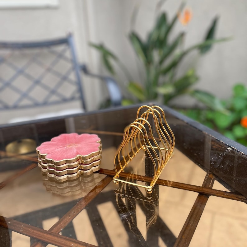 Puede incluir: Un juego de cuatro posavasos de color rosa en forma de flor con ribete dorado apilados uno encima del otro. Los posavasos est&aacute;n sobre una mesa de vidrio junto a un portavasos de metal dorado.