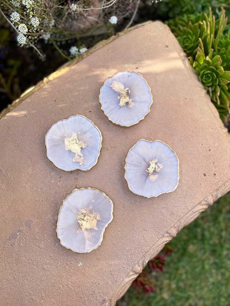 May include: Four light purple resin coasters with gold flecks and a gold rim. The coasters are on a brown surface.