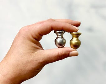 Puede incluir: Dos pequeños remates, uno plateado y otro dorado, se sostienen en una mano. Ambos remates son redondos y tienen una pequeña parte superior decorativa.