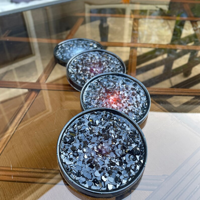 May include: Four black coasters with a glittery, iridescent finish. The coasters have a black rim and a clear, resin-like center filled with black glitter. The glitter reflects light, creating a shimmering effect.