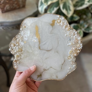 May include: A decorative, white resin tray with a gold trim and accents. The tray is adorned with gold floral embellishments, pearls, and gold leaf details. The tray is held by a hand.