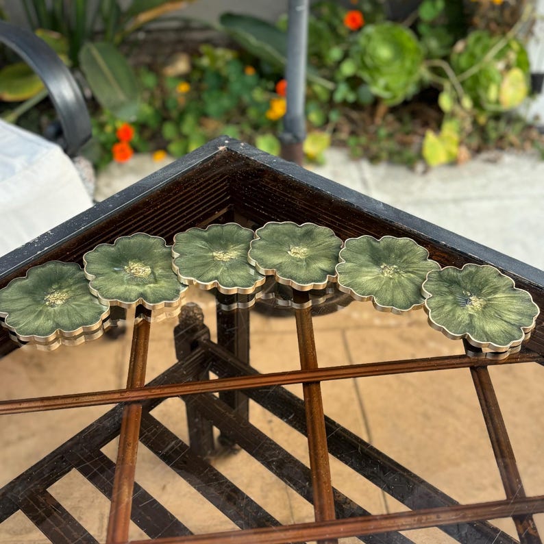 Peut inclure: Six sous-verres en r&eacute;sine verte et dor&eacute;e avec un motif floral. Les sous-verres sont dispos&eacute;s en rang&eacute;e sur une table en verre.