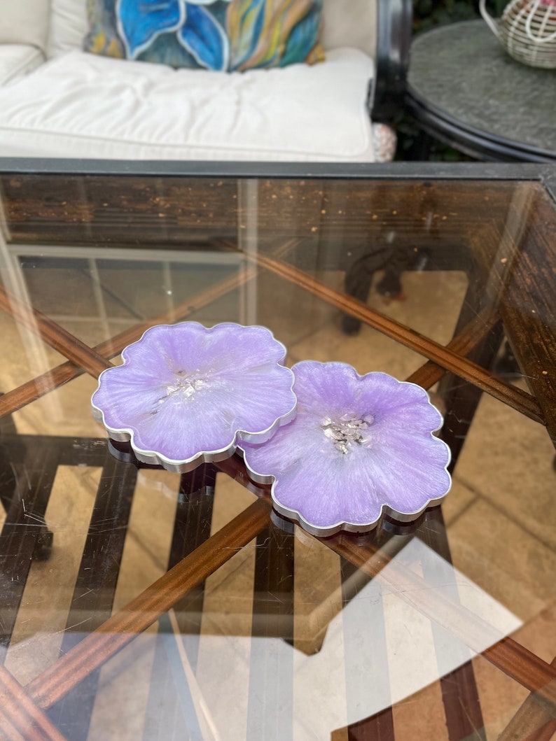 Op de afbeelding: Twee paarse onderzetters van hars met een zilveren glitteraccent. De onderzetters zijn bloemvormig en hebben een zilveren rand.