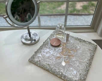 May include: A silver-toned, lighted makeup mirror stands on a white vanity table. A decorative tray with a white and gold design, surrounded by clear crystals, holds a perfume bottle, makeup, and a lipstick. The vanity is near a window.