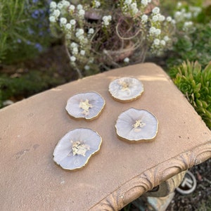 May include: Four white and gold resin coasters with a unique agate design. The coasters are round and have a gold rim.