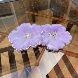 Op de afbeelding: Twee paarse harsonderzetters met een bloemachtig ontwerp en zilveren glitteraccenten. De onderzetters staan op een glazen tafel met een houten basis.