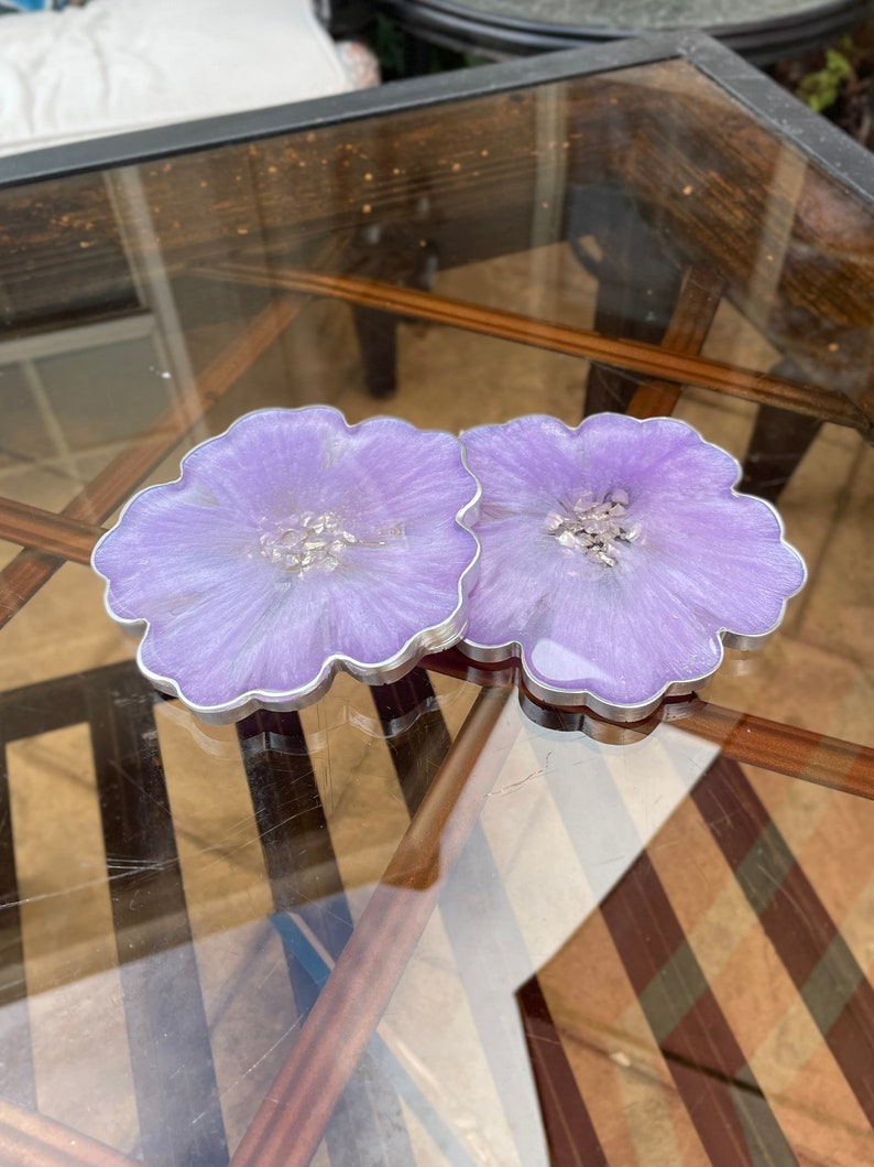Op de afbeelding: Twee paarse harsonderzetters met een bloemachtig ontwerp en zilveren glitteraccenten. De onderzetters staan op een glazen tafel met een houten basis.