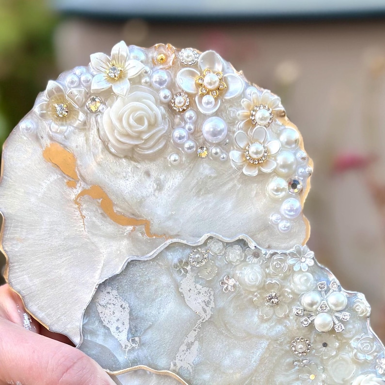 May include: Two decorative resin coasters. The coasters are white with gold accents and are adorned with white flowers, pearls, and rhinestones. The edges are trimmed with gold. The coasters have a glossy finish and a unique, artistic design.