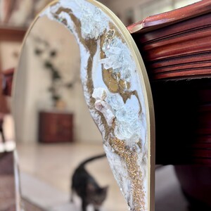 May include: An arched mirror featuring a decorative border embellished with white resin, gold paint, and clear crystalline rock fragments. The frame has a smooth gold finish, and the reflective surface shows a blurred interior space.