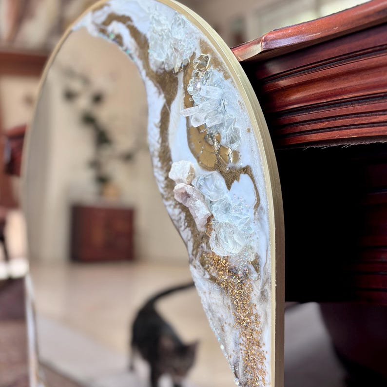 May include: An arched mirror featuring a decorative border embellished with white resin, gold paint, and clear crystalline rock fragments. The frame has a smooth gold finish, and the reflective surface shows a blurred interior space.