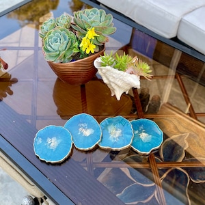 May include: Four blue and gold resin coasters with a white marbled design. The coasters are on a glass table top with a potted succulent plant and a seashell.