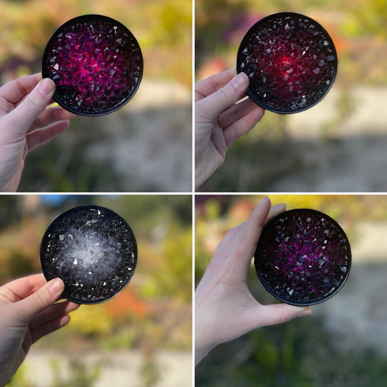 May include: Four round black coasters with a clear resin top filled with iridescent purple and white glitter. The coasters are being held by a hand.