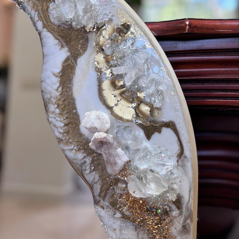 May include: A decorative resin art piece featuring a swirling white and gold marbled pattern. The surface is embedded with clear quartz crystals, small white stones, and fine gold glitter, creating a textured, layered appearance with metallic accents.