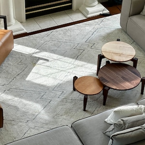 May include: Three round wooden nesting tables with a light wood top, a dark wood middle, and a light wood bottom. The tables are on a white rug with a geometric pattern.
