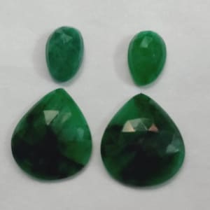 May include: Four faceted green gemstones. Two are teardrop-shaped, and two are oval-shaped. The gemstones have a dark green color with lighter green facets. The gemstones are arranged in a square formation.