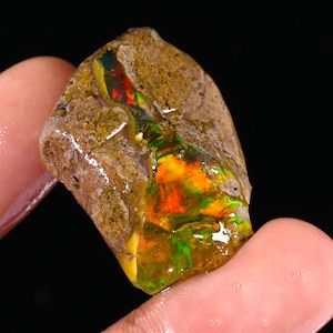 Puede incluir: Una gema de ópalo en bruto, sin cortar, con una matriz marrón. El ópalo muestra colores internos vibrantes de rojo, naranja y verde. La gema se sostiene entre los dedos, mostrando su forma natural y en bruto.