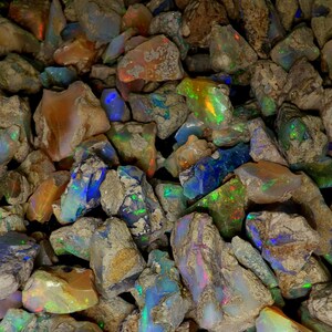 Op de afbeelding: Een close-up van een stapel ruwe, onbewerkte opaalstenen. De stenen zijn in verschillende tinten bruin, grijs en blauw met iriserende kleurvlekken.