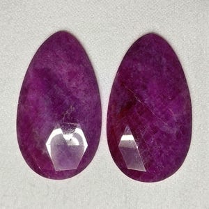 Könnte beinhalten: Zwei facettierte, tropfenförmige Edelsteine in tiefem Magenta. Die Edelsteine haben eine polierte Oberfläche mit inneren Adern und eine große, geometrische Facette nahe der Basis. Die Steine werden auf weißem Hintergrund präsentiert.